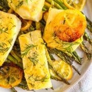 4 pieces of sea bass on a bed of asparagus on a white plate and garnished with lemons and dill.