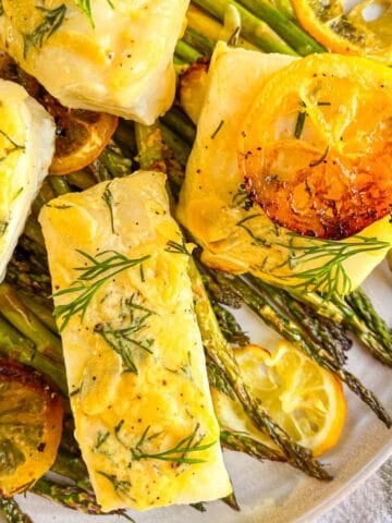 4 pieces of sea bass on a bed of asparagus on a white plate and garnished with lemons and dill.