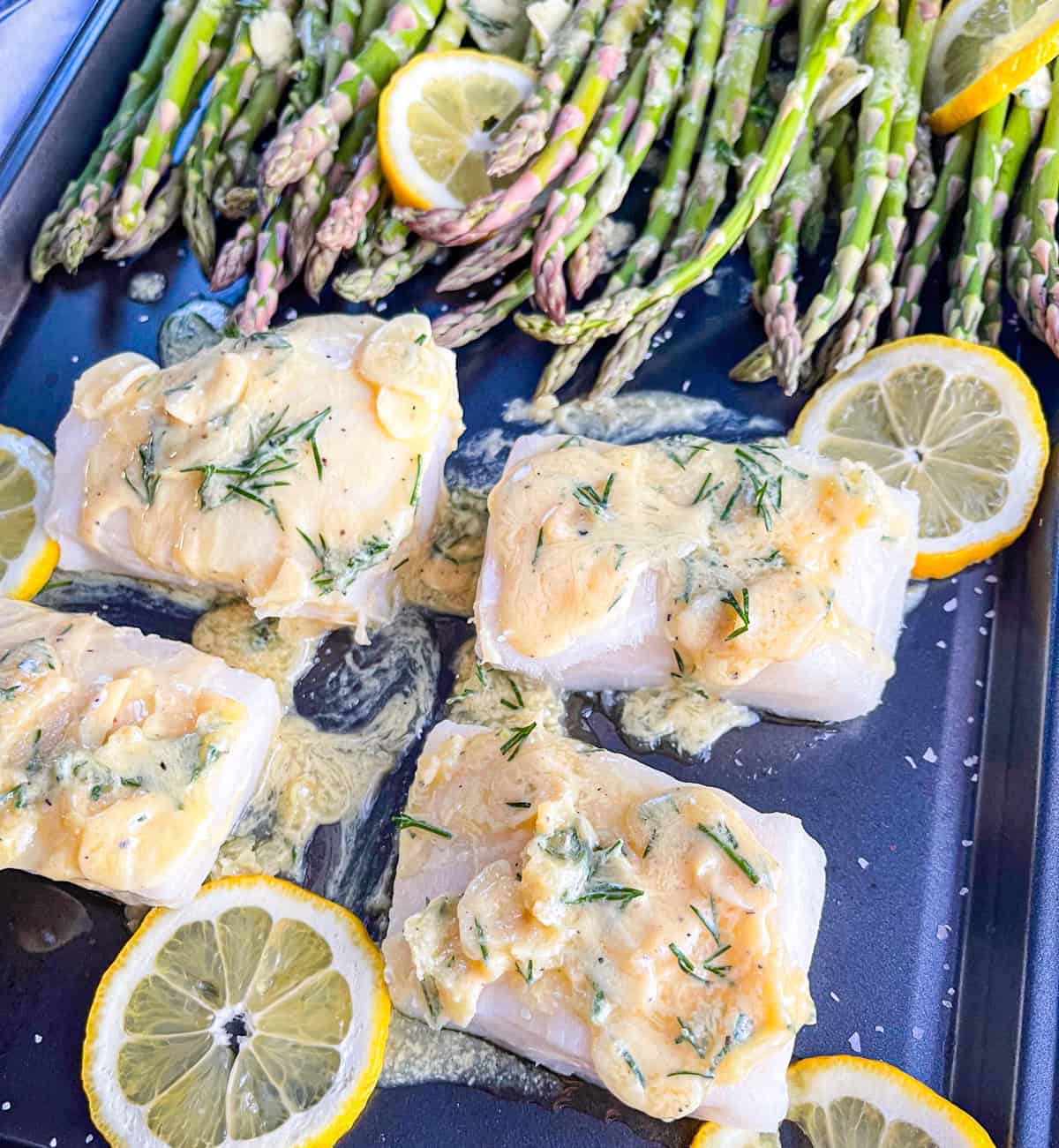 four fish fillets on a sheet tray with garlic lemon butter sauce and a row of asparagus.
