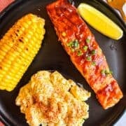Grilled bbq salmon on a black plate with potato salad and corn.