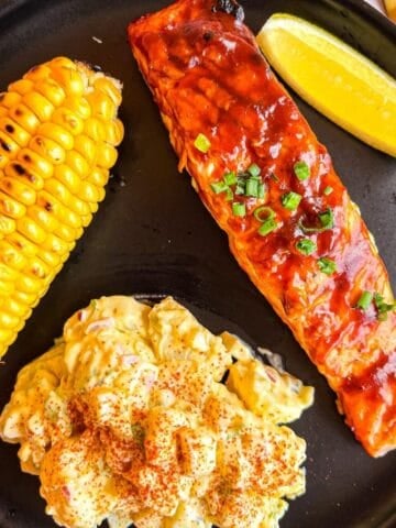 Grilled bbq salmon on a black plate with potato salad and corn.