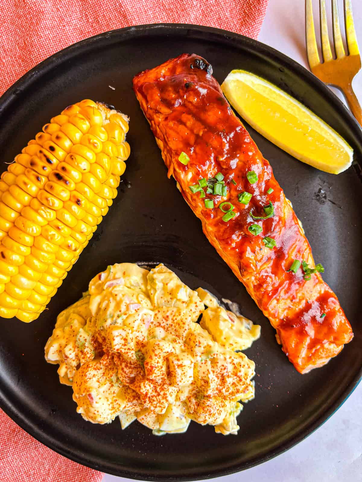 Grilled bbq salmon on a black plate with potato salad and corn.