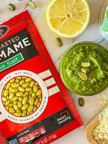 Edamame Pesto on a table with edamame around next to basil, parmesan and lemon.