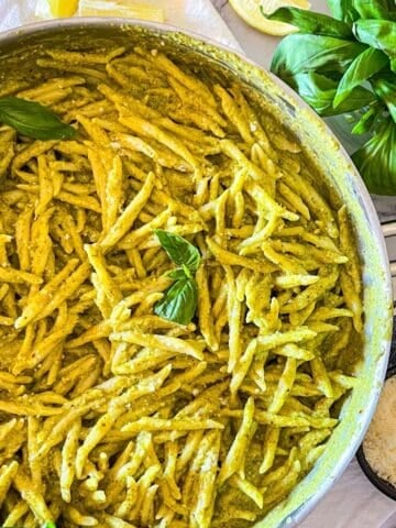 Pesto pasta in a skillet with basil and cheese next to it.