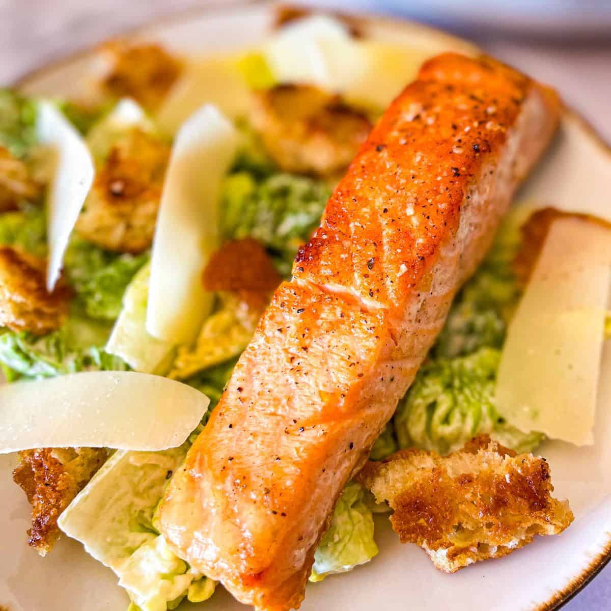 Salmon Caesar Salad garnished with large shreds of parmesan and homemade croutons.